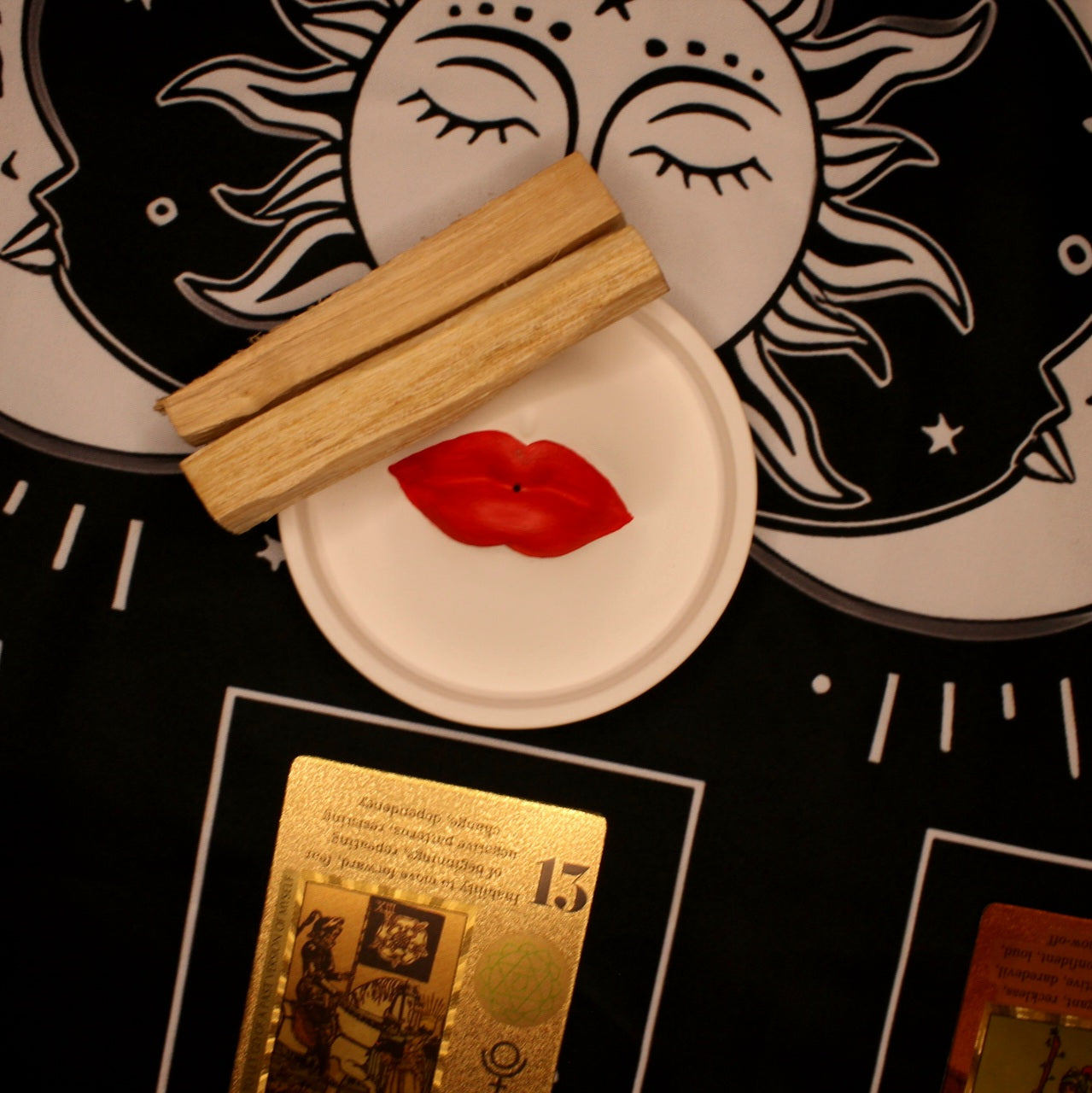 Kawałki drewna palo santo oraz podstawka w kształcie ust na tle mistycznej tkaniny z symbolami słońca i księżyca.