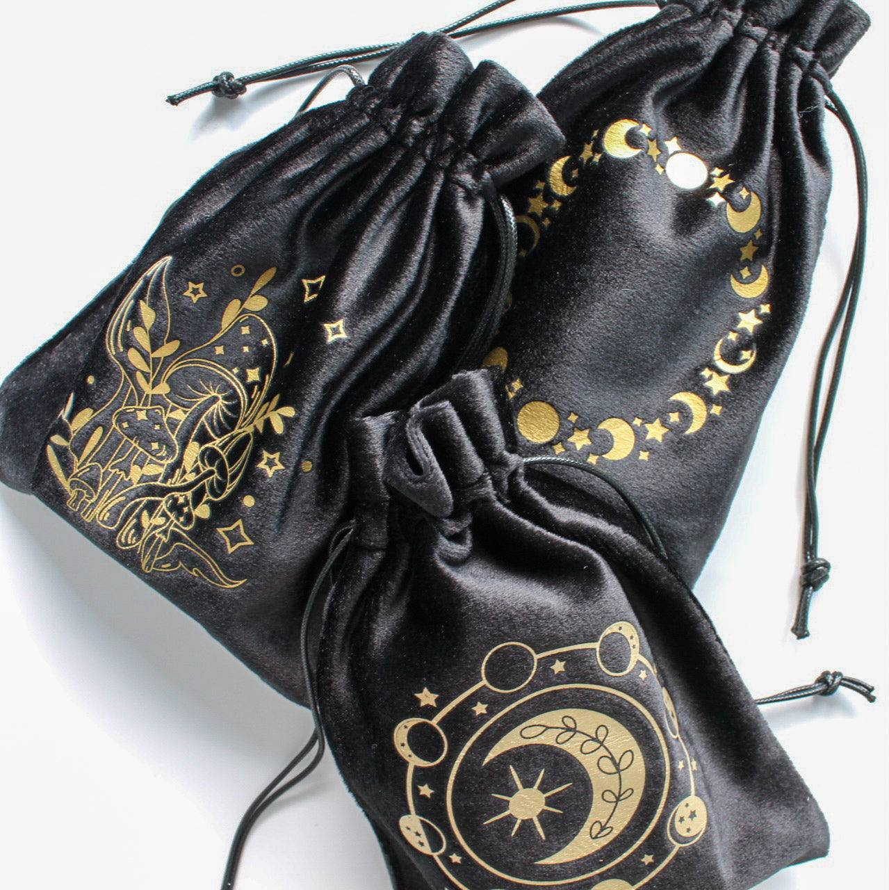 Trzy czarne woreczki o aksamitnej fakturze, każdy z unikalnym złotym wzorem (grzyby, księżycowe fazy, motyw kosmiczny), rozłożone na białym tle.