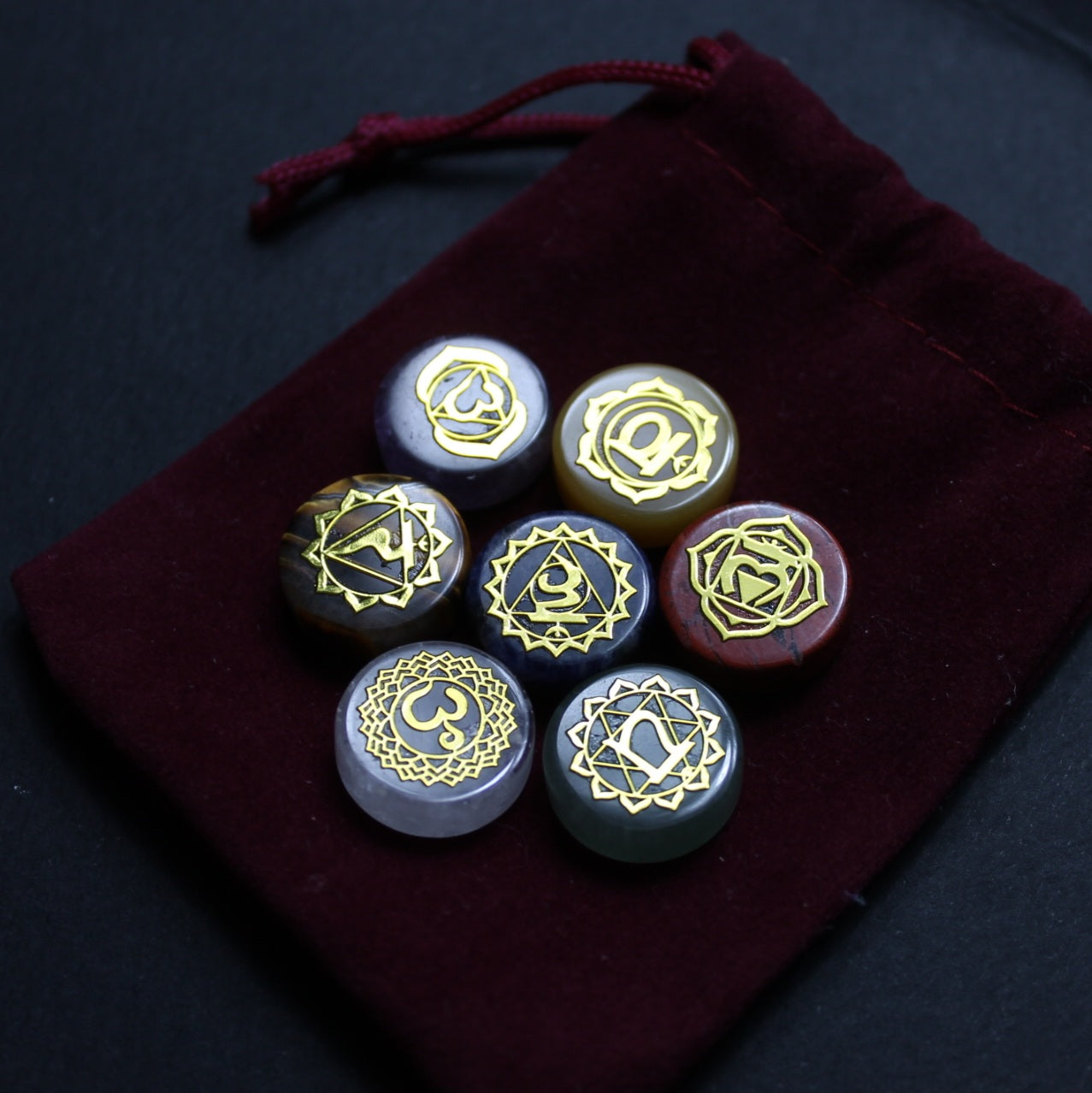 Siedem kolorowych kamieni czakr ze złotymi symbolami sanskryckim ułożonych na bordowym aksamitnym woreczku ze sznurkiem, sfotografowanych na ciemnym tle