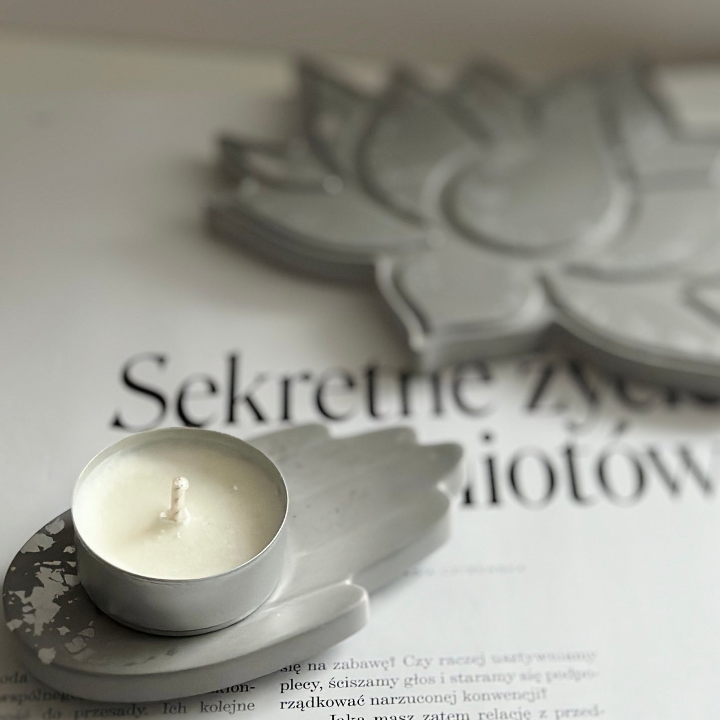Zbliżenie na białą świecę umieszczoną na świeczniku w kształcie dłoni. W tle widać szarą tackę w formie lotosu oraz fragment otwartego magazynu.