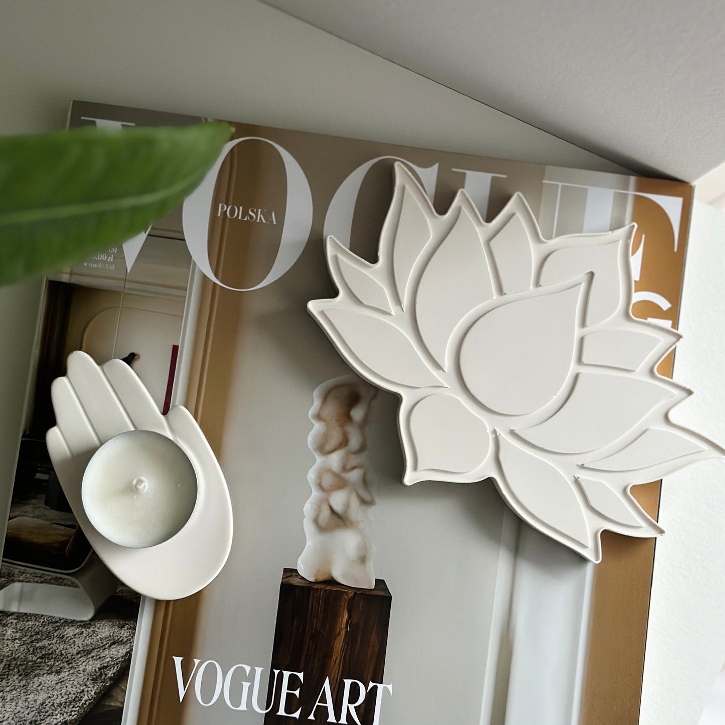Kremowa tacka w kształcie kwiatu lotosu i świecznik w formie dłoni, ustawione na magazynie "Vogue Art". Widać również zielony liść jako element dekoracyjny.