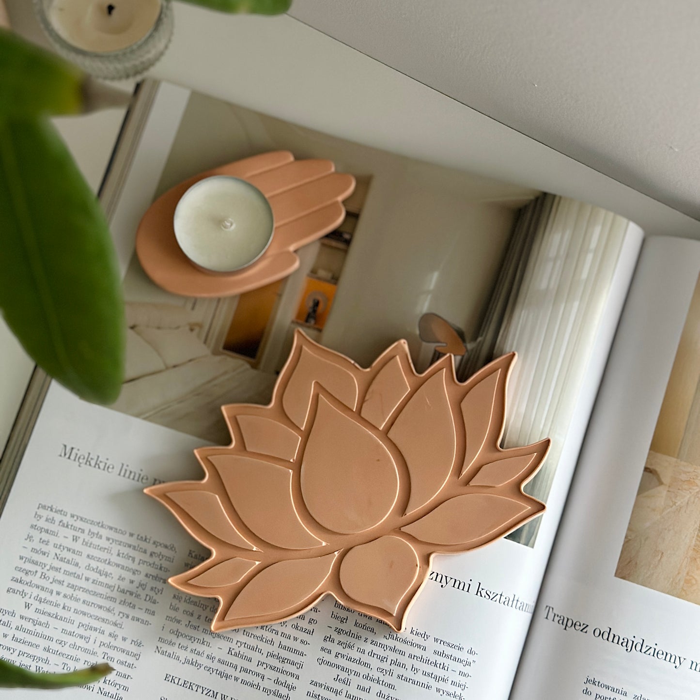 Beżowa tacka w kształcie lotosu, obok świecznik w kształcie dłoni. Tło stanowi otwarty magazyn z aranżacją wnętrz.