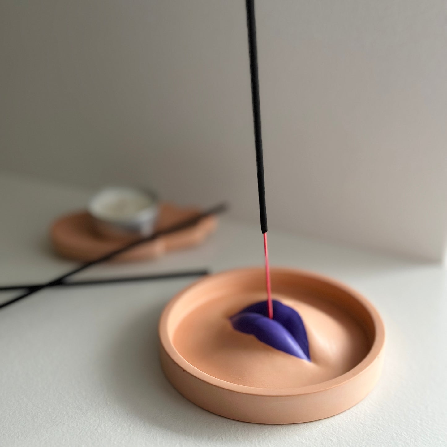 Okrągła, brzoskwiniowa podstawka z fioletowym detalem w kształcie ust, służąca jako podstawka na kadzidło.