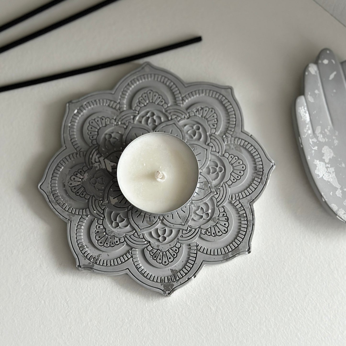 Szara podstawka w kształcie mandali, ozdobiona misternymi, symetrycznymi wzorami, na której znajduje się biała świeczka. Obok leży ozdobna tacka w formie dłoni oraz czarne patyczki zapachowe.