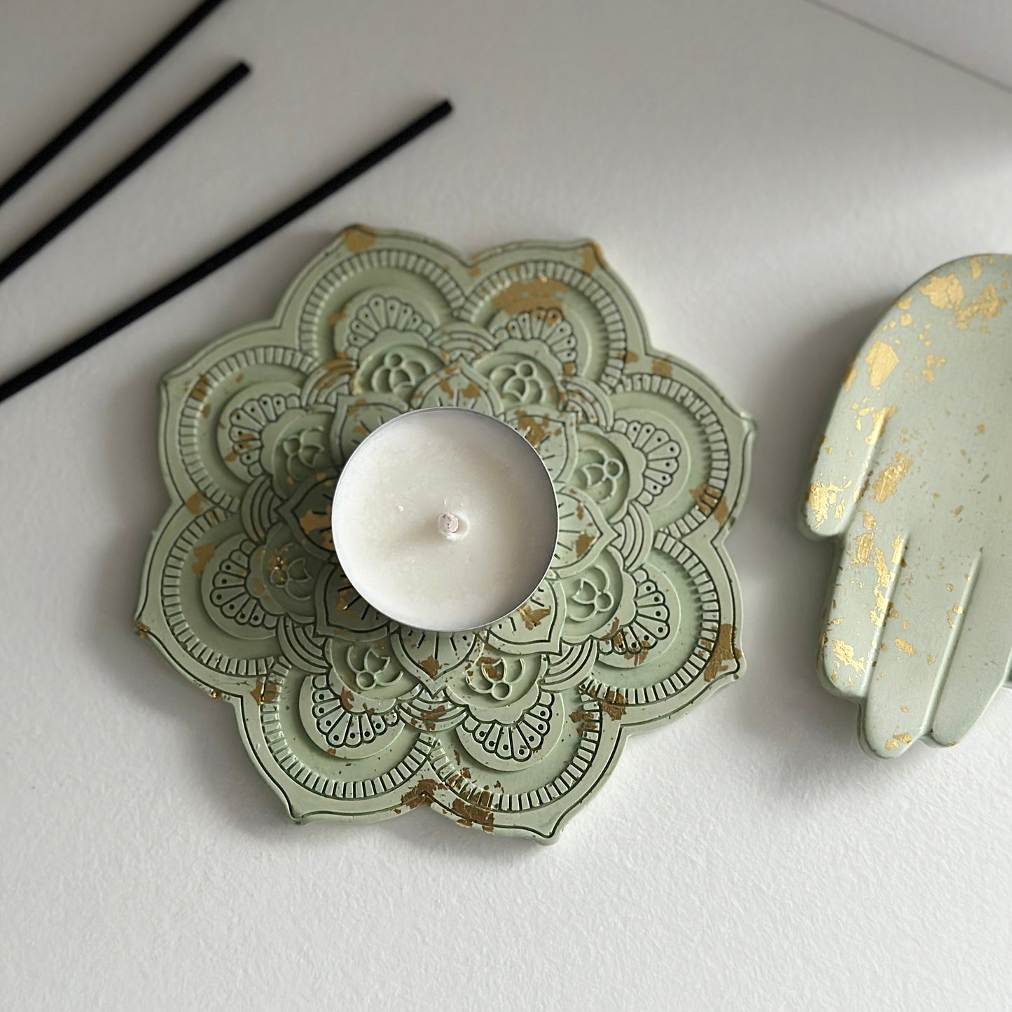 Zielona podstawka mandalowa z delikatnymi złotymi akcentami, trzymająca białą świeczkę. Obok widoczny jest świecznik w kształcie dłoni z podobnym złotym zdobieniem.
