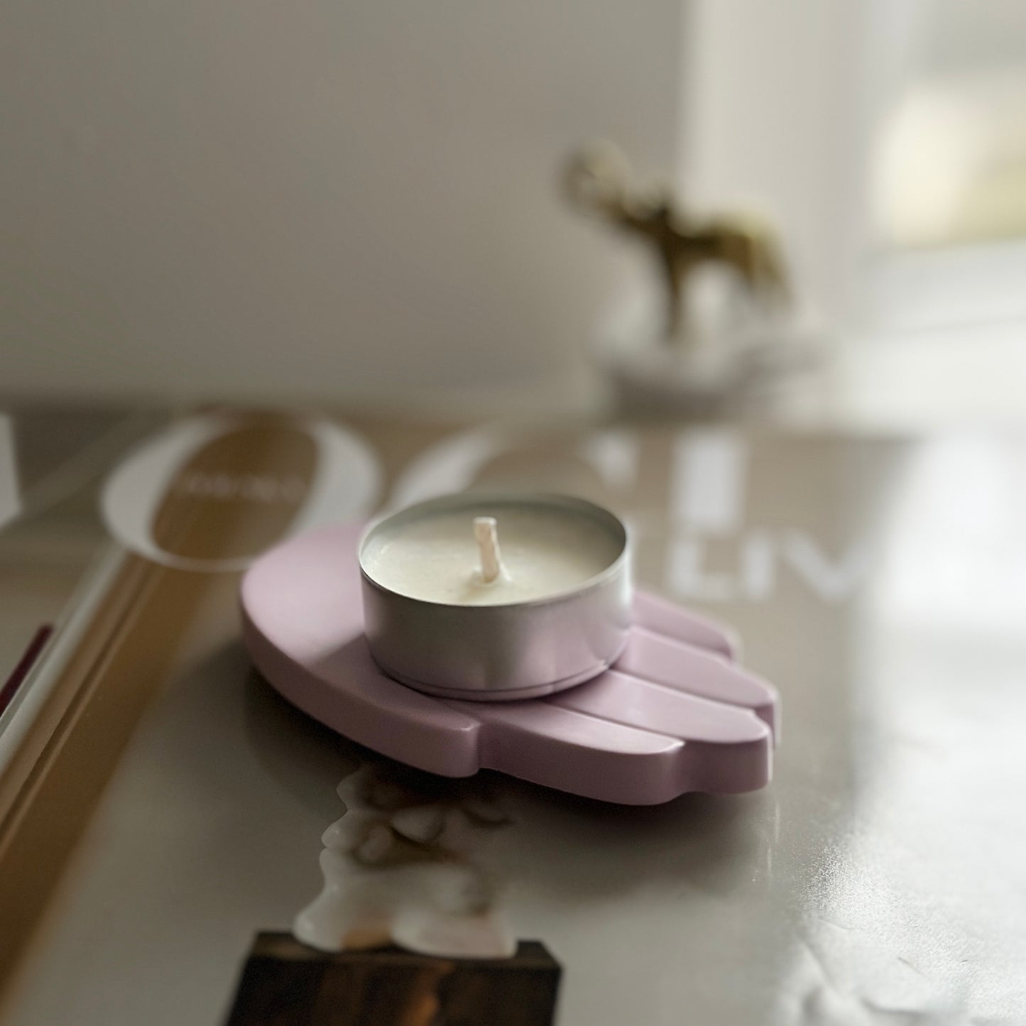 Świecznik w kształcie dłoni w jasnoróżowym kolorze, trzymający białą świeczkę typu tealight. Ułożony na magazynie "Vogue", w tle widoczna rozmyta figurka złotego słonia.