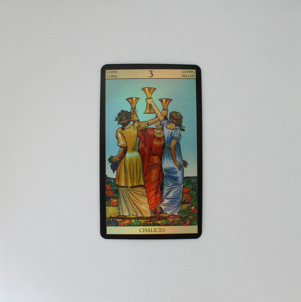 Karta tarota „3 of Chalices” przedstawiająca trzy kobiety, które stoją tyłem do obserwatora, wznosząc złote kielichy w geście toastu, ubrane w różnobarwne szaty.