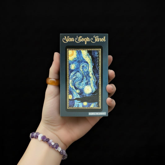 Dłoń trzymająca pudełko talii kart "Van Gogh Tarot" z grafiką inspirowaną "Gwiazdną nocą", osoba ma na nadgarstku bransoletę z fioletowych i białych kamieni.