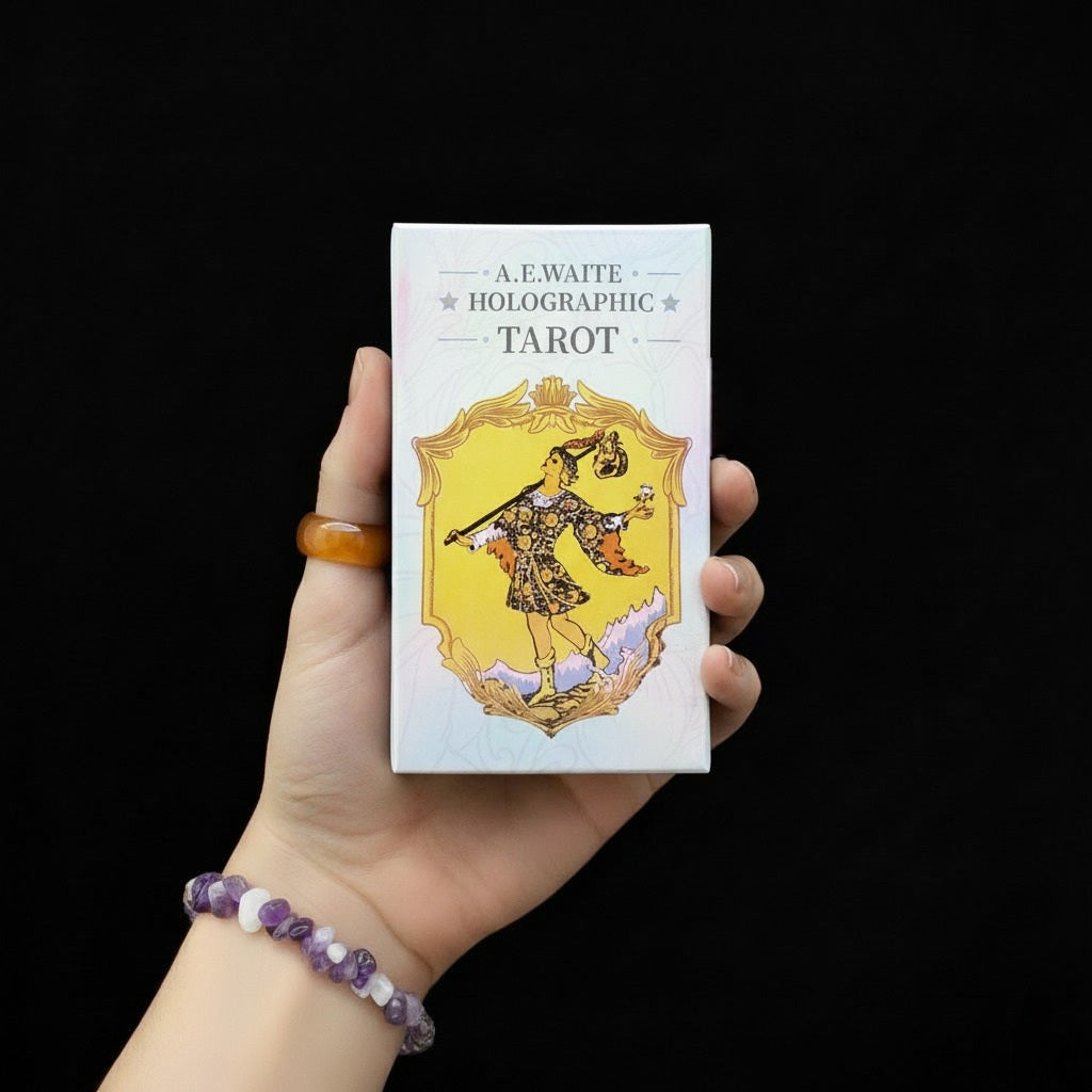 Dłoń trzymająca pudełko talii kart "A.E. Waite Holographic Tarot" z białym tłem i postacią w ozdobnej złotej ramie, osoba ma na nadgarstku bransoletę z fioletowych i białych kamieni.