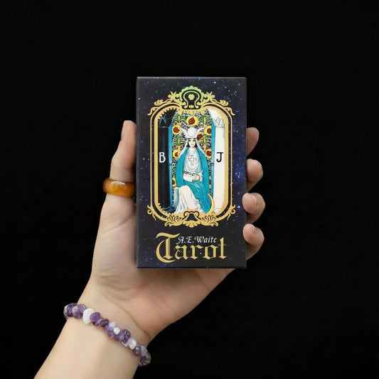 Dłoń trzymająca pudełko talii kart "A.E. Waite Tarot" z ciemnym gwiaździstym tłem i kartą Arcykapłanki w ozdobnej złotej ramie, osoba ma na nadgarstku bransoletę z fioletowych i białych kamieni.