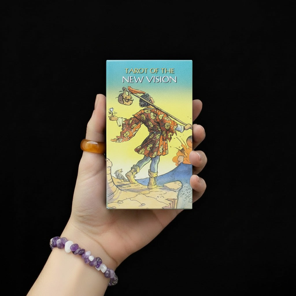 Dłoń trzymająca pudełko talii kart "Tarot of the New Vision" z ilustracją postaci trzymającej miecz na tle turkusowo-żółtego nieba, osoba ma na nadgarstku bransoletę z fioletowych i białych kamieni.