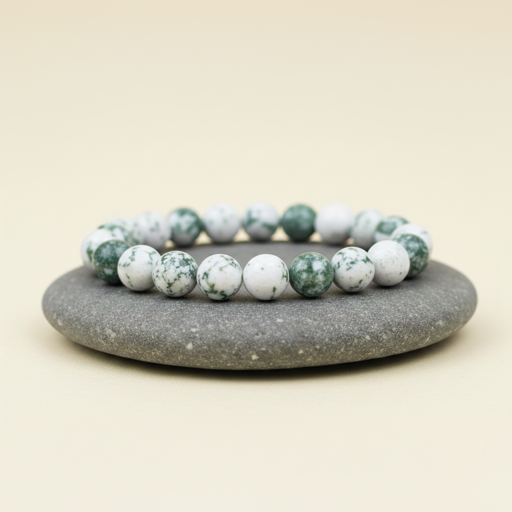 Bransoletka z okrągłych kamieni w kolorze białym z zielonymi plamkami, z metalowymi zdobieniami, ułożona na szarym kamieniu na beżowym tle.