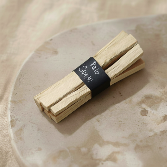 Pęczek drewna Palo Santo przewiązany czarną opaską z białym napisem "Palo Santo", ułożony na jasnej kamiennej lub ceramicznej płytce na beżowym tle