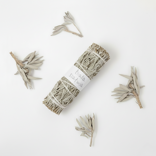 Pęczek białej szałwii przewiązany sznurkiem z etykietą "biała szałwia", otoczony luźnymi suszonymi liśćmi szałwii ułożonymi na białym tle