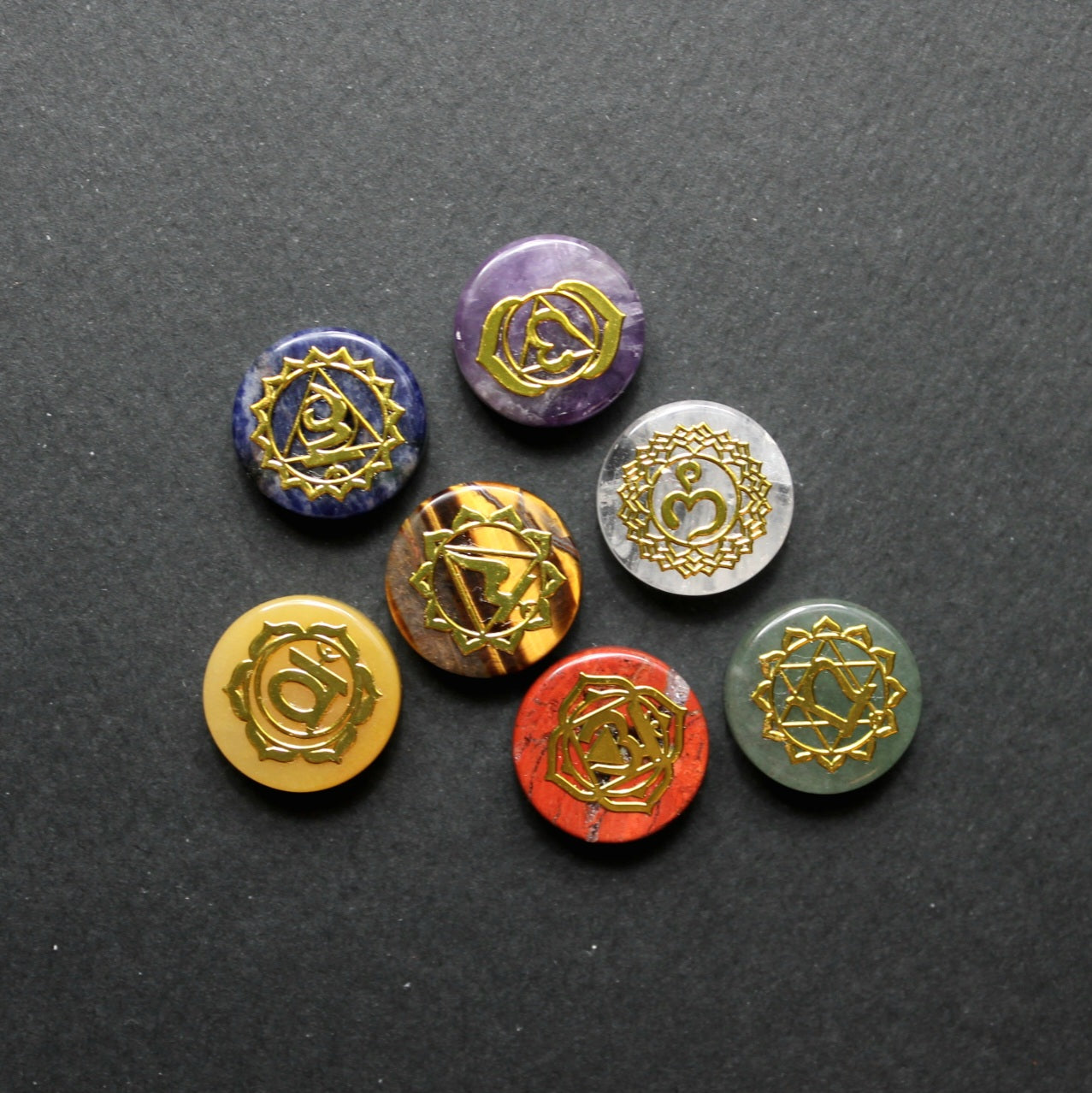 Siedem kamieni czakr ułożonych w okrąg na ciemnoszarym teksturowanym tle - widoczne kolory: fioletowy, żółty, pomarańczowy, biały, niebieski, zielony i czerwony, każdy z wygrawerowanym złotym symbolem czakry