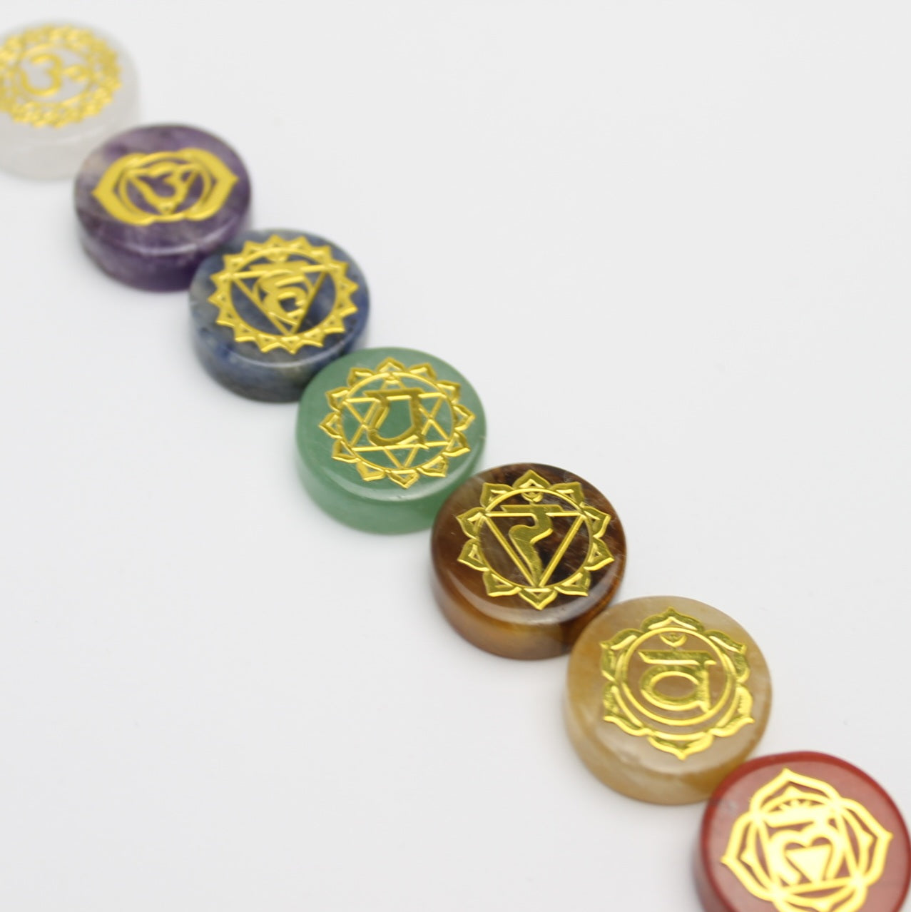 Siedem kamieni czakr ułożonych po przekątnej od lewego górnego do prawego dolnego rogu na białym tle, ukazujących pełne spektrum kolorów od fioletu do czerwieni