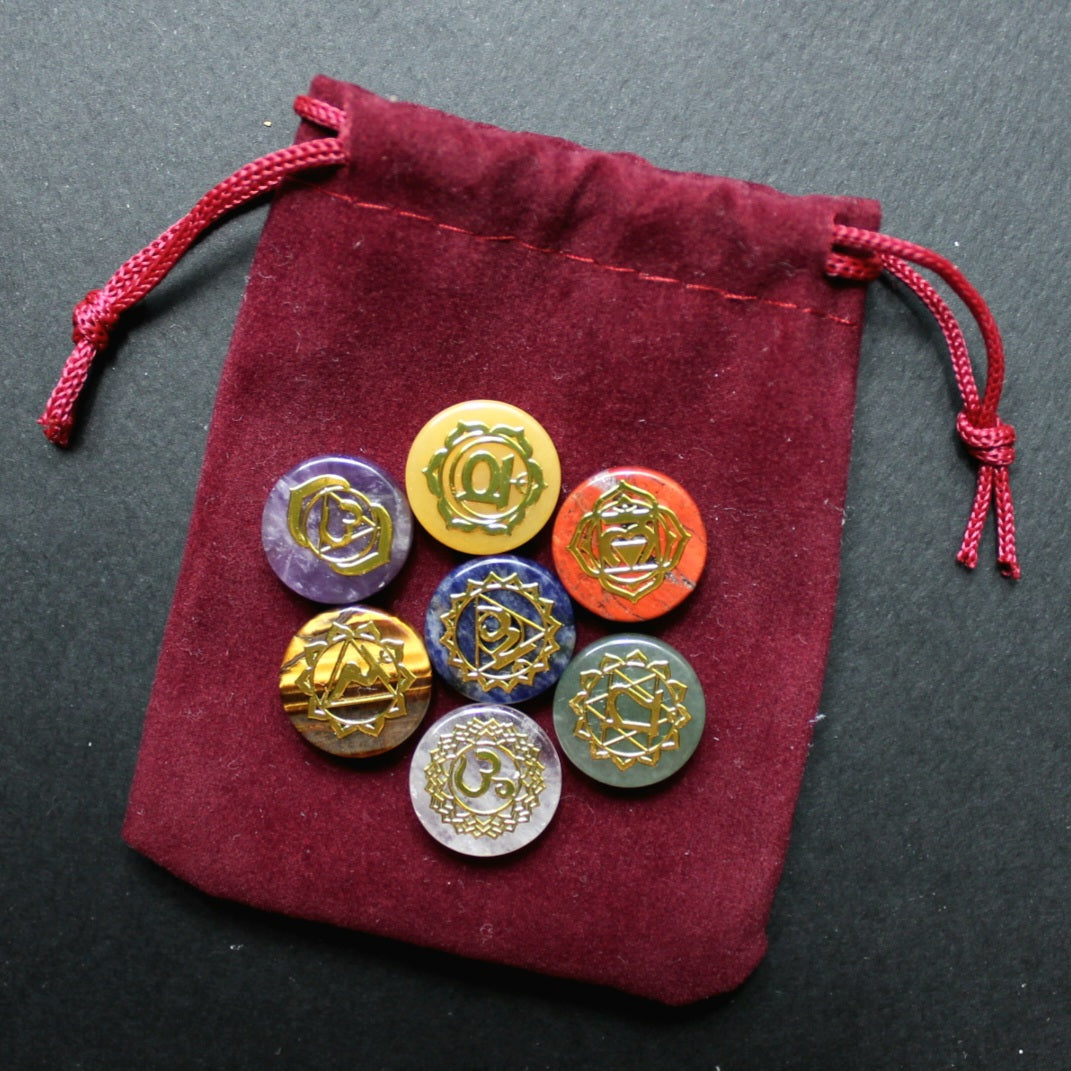 Siedem kamieni czakr ze złotymi symbolami leżących na bordowym aksamitnym woreczku ze sznurkiem, sfotografowanych na ciemnoszarym tle