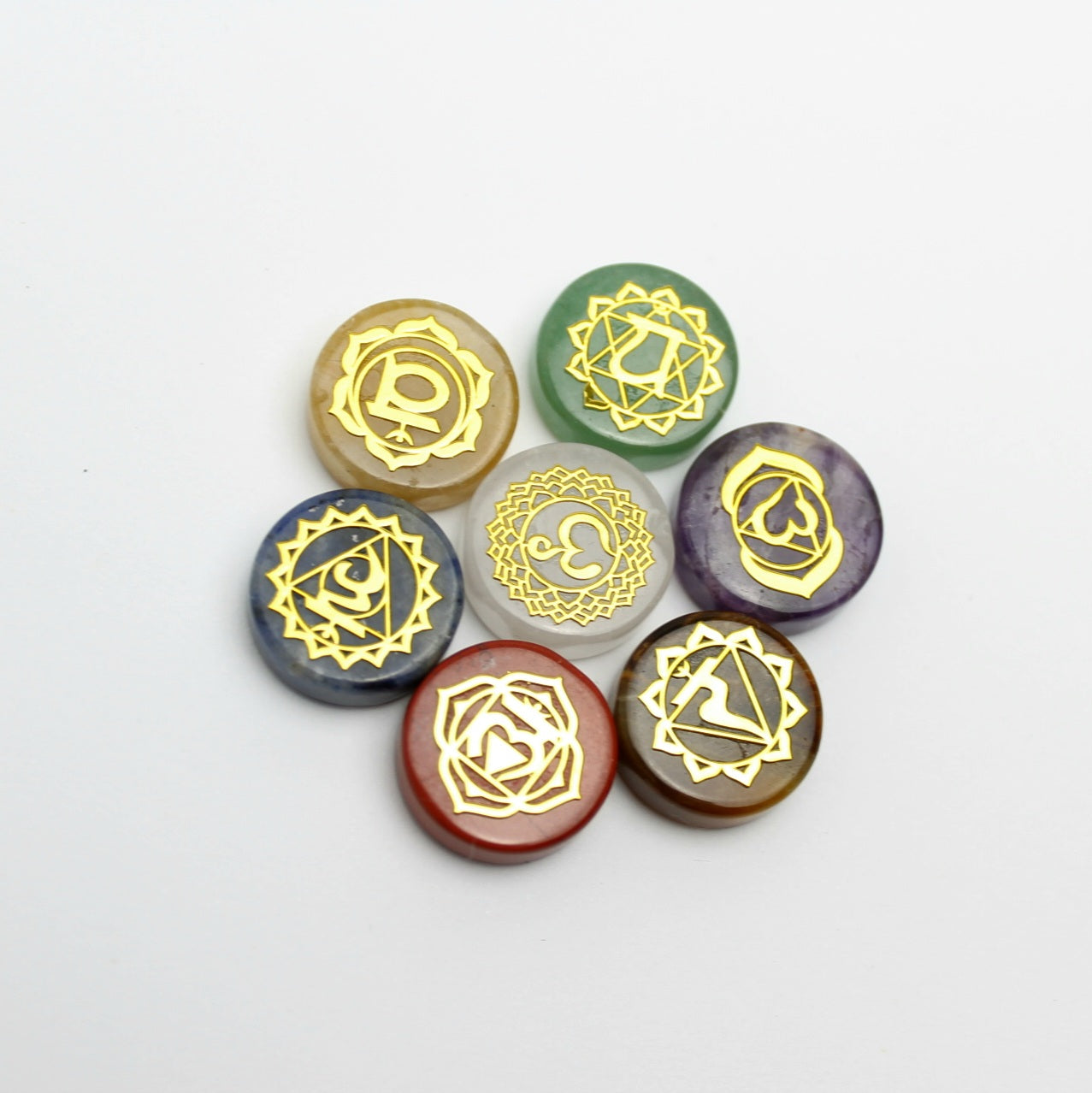 Siedem kamieni czakr ułożonych w okrągły wzór na białym tle, pokazujących szczegółowe złote grawerowane symbole sanskryckie na kolorowych kamieniach
